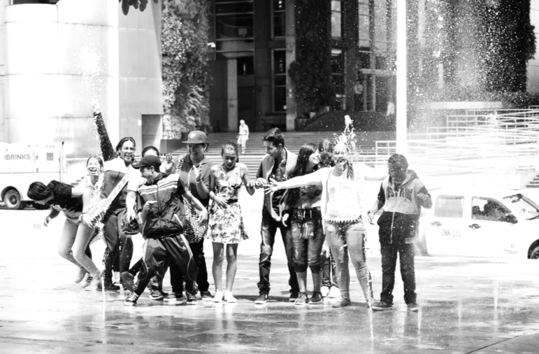 A black and white picture of a group of people, mostly young people that are playing around a water fountain.