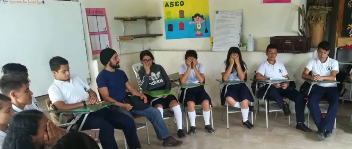 A group of students sitting in a semi-circle, and one teacher. Some of them have their hands over their faces, others have their eyes closed.