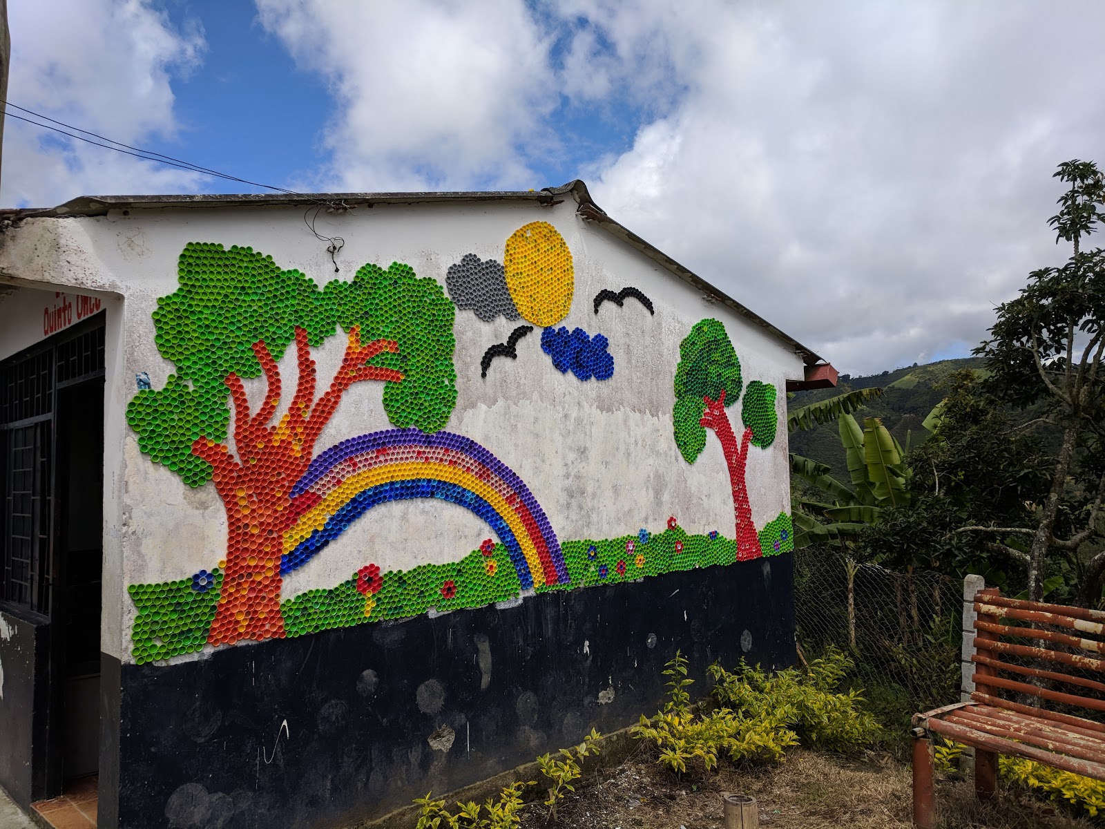 Mural on a school of trees, sun, clouds, birds and a rainbow made of brightly coloiured plastic bottle caps
