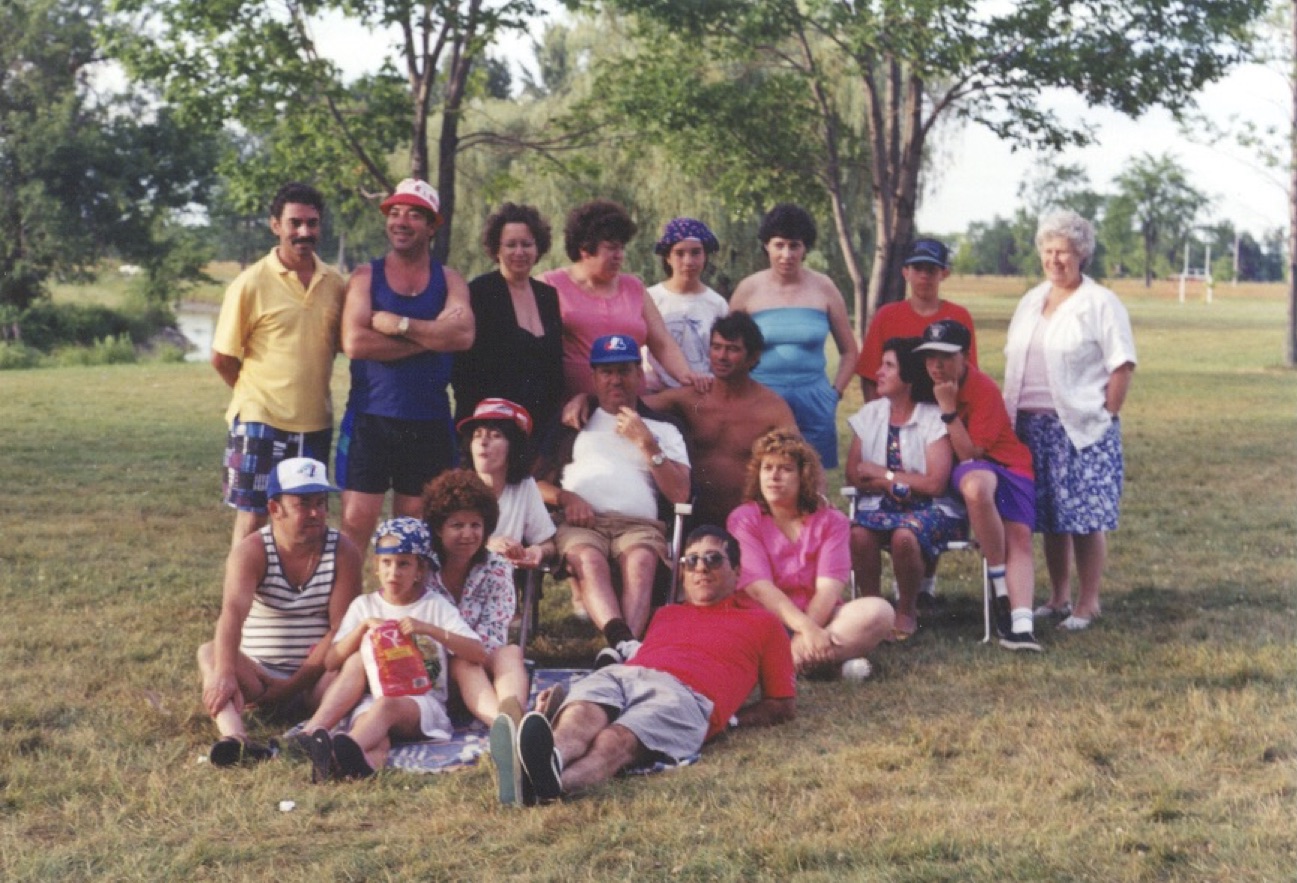 A large group of people, including one small child, sit and stand on the grass near some trees. Many are smiling for the camera. They are dressed for warm weather, with many wearing shorts and T-shirts or tank tops.
