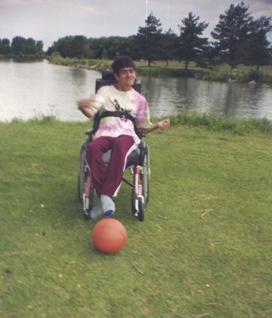 A young man sits in a wheelchair on some grass in front of a pond surrounded by trees, with a basketball lying on the ground in front of him. He is smiling, and his right arm appears to be in motion. He has short brown hair and wears burgundy track pants, grey socks and a pink and yellow tie-dyed T-shirt.