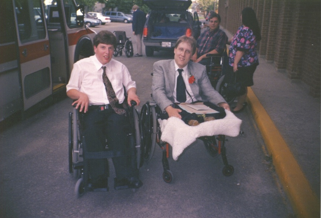 Two young men in wheelchairs sit side by side at the edge of the street between a small bus and a brick building. They are dressed formally, both wearing ties, with one in a blazer, and are posing and smiling for the camera. In the background there is a man standing beside a wheelchair and an SUV, as well as two people standing on the street directly behind them. 