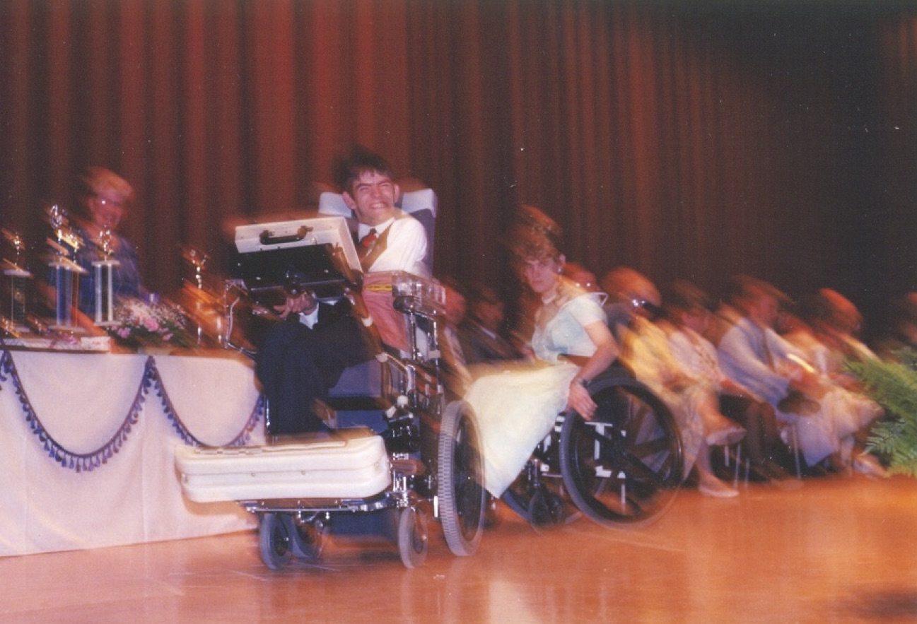 A blurry photo shows a young man in a power wheelchair who appears to be moving across a stage. He is beside a table full of trophies, with a number of people sitting behind him on the stage including one woman in a wheelchair.
