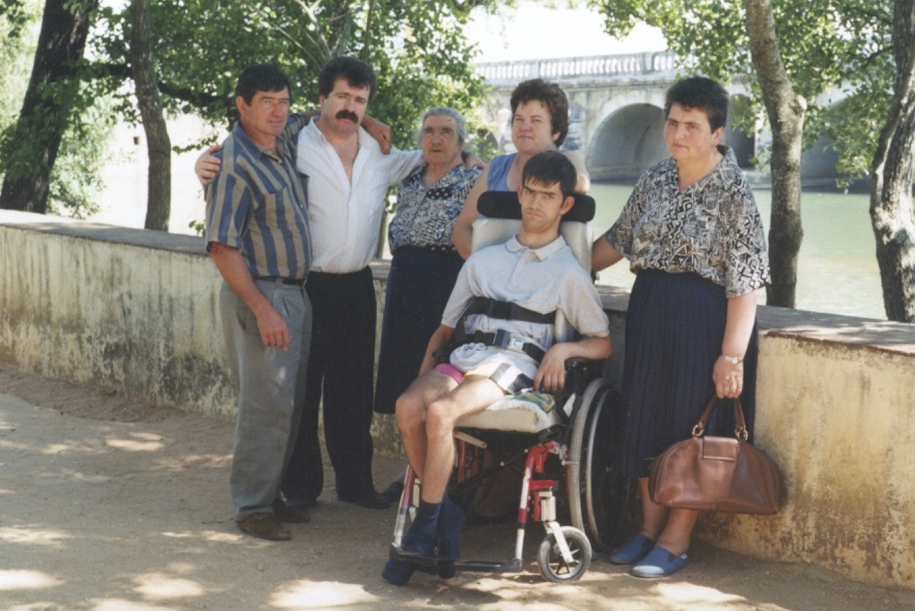 A group of 6 people, including 3 men and 3 women, pose in front of a river under some trees. A bridge appears in the background. One man sits in a wheelchair.