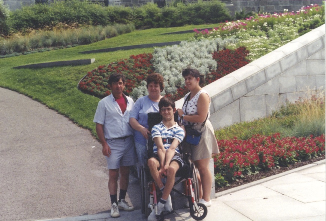 A group of 4 people pose in front of a large, landscaped garden of grass and flowers. One man and 2 women stand behind a young man sitting in a wheelchair.