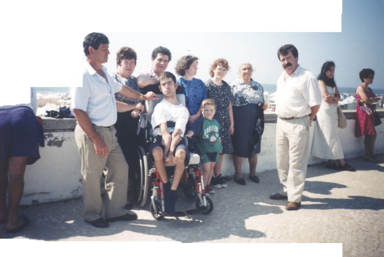 A group of people, including a child and a man in a wheelchair, pose in the sun on a road overlooking what appears to be some water and buildings. 