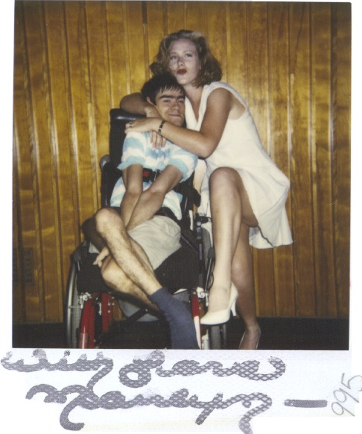 An autographed polaroid (autograph illegible) showing a young man in a wheelchair being embraced by a woman in a short white dress and high heels, who stands on one foot, while lifting the other leg toward the camera. 
