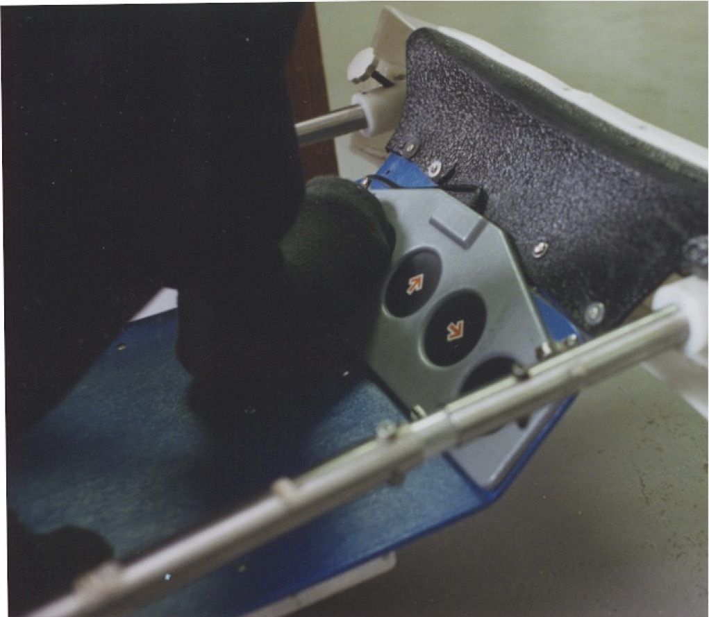 Close-up view of someone's foot controlling the large buttons of a communication aid, which are mounted on a foot rest, two of which are visible and are labelled with an up arrow and a right arrow respectively.