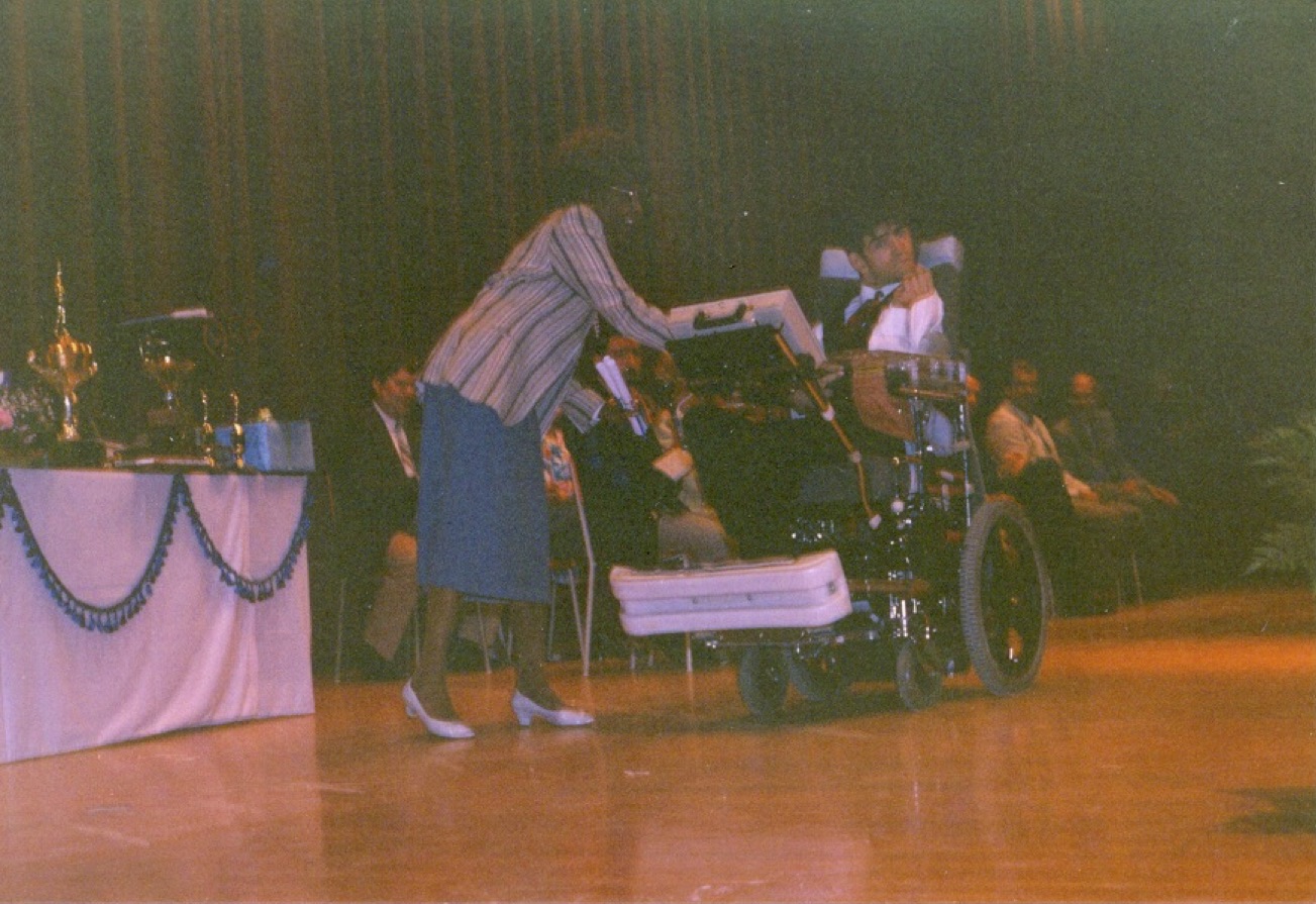 A young man sits in a power wheelchair on a stage.  A woman stands beside him and appears to be handing him something.
