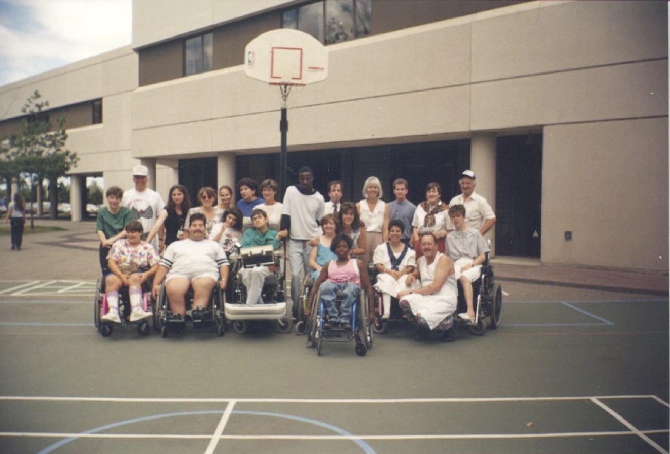 A large group of people pose on an outdoor basketball court, under a basket. Many of them are in wheelchairs in the front row, while others stand behind.