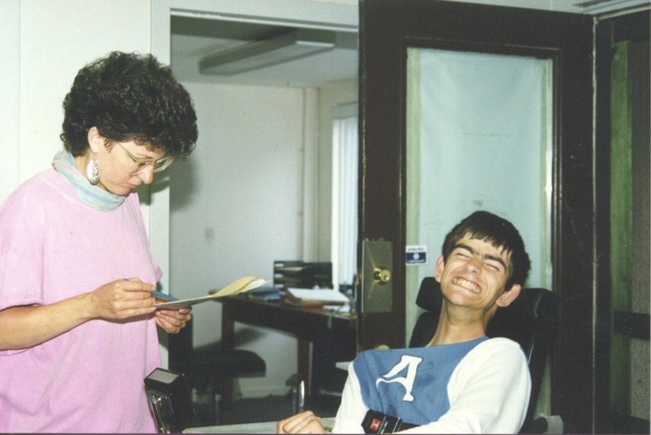 A young man in a wheelchair sits smiling in front of a woman who stands, holding and reading some papers. They appear to be in an office.